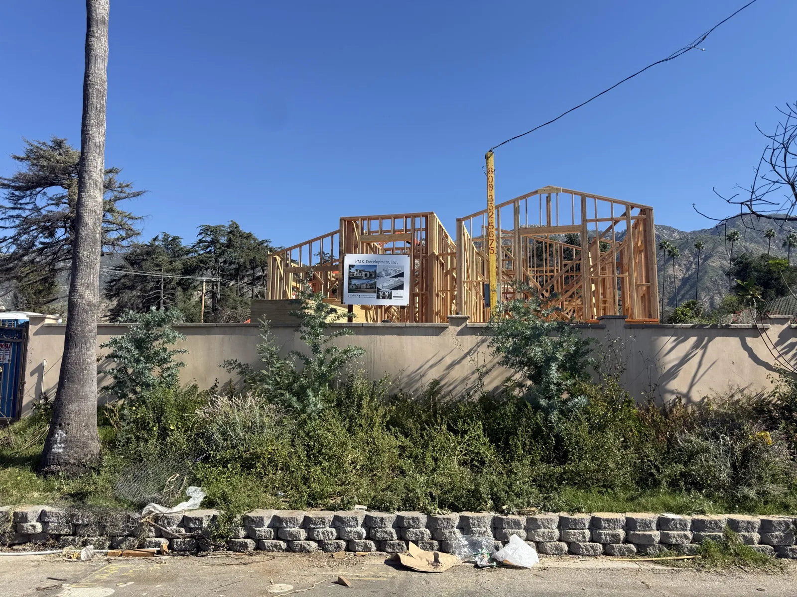 Wide street view of full first-floor stud framing with the PMK jobsite sign and the San Gabriel mountains behind.