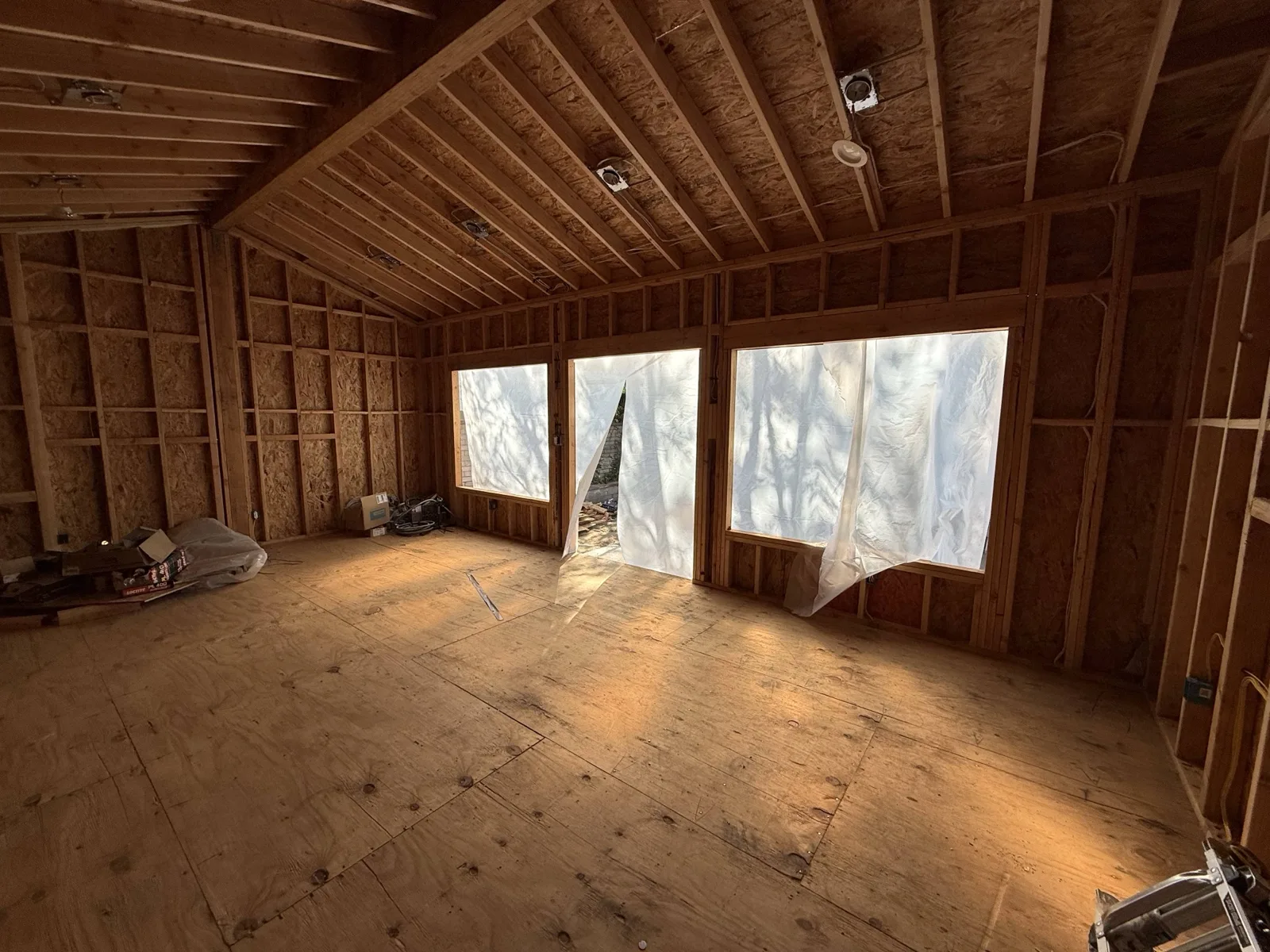 Framed vaulted interior with window and door openings sheeted in plastic, recessed-light boxes wired overhead.