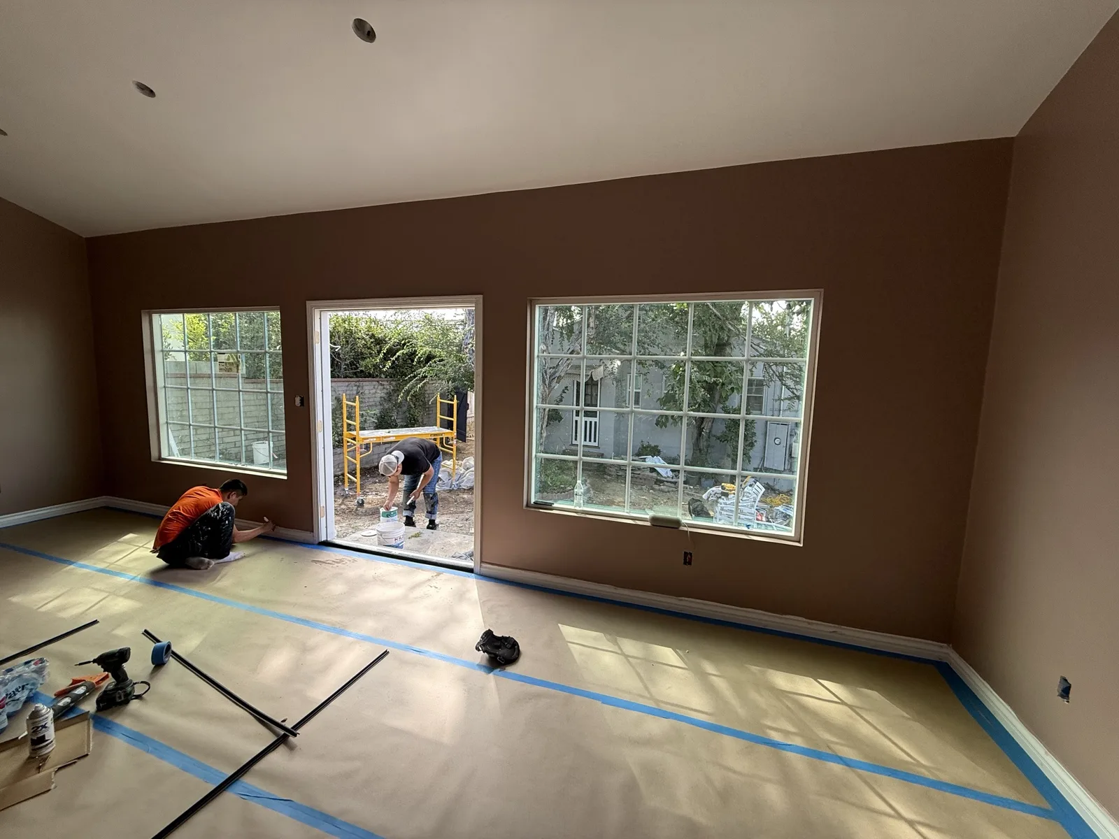 Painters detailing the baseboards in the finished interior, floor protection down and blue tape lines in place.