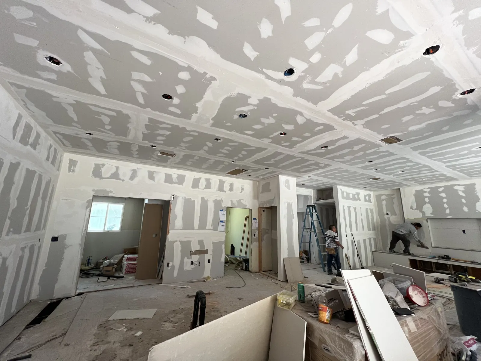 Crew taping and mudding drywall seams across the ceiling and walls of the main living area, with recessed lighting openings cut in, at PMK's addition-and-renovation project in La Cañada Flintridge.