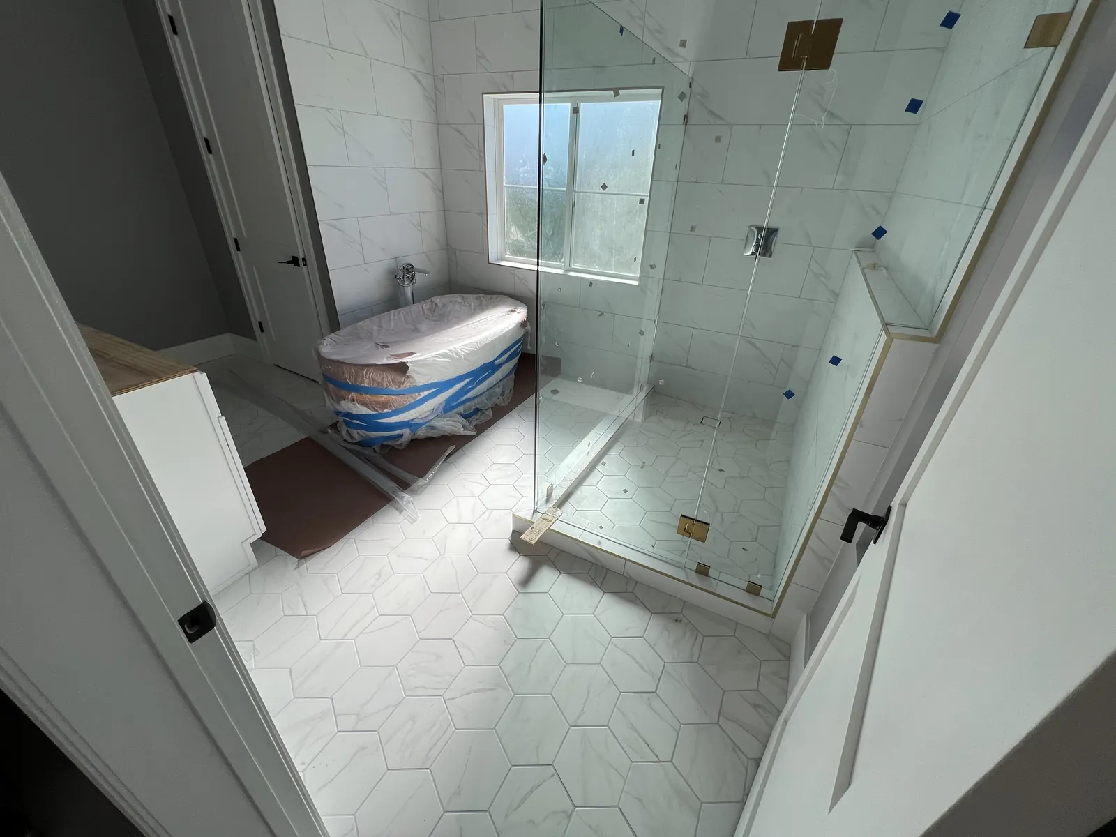 Primary bathroom mid-finish with hexagonal marble-look floor tile, a frameless glass shower enclosure, and the freestanding tub still in protective wrap at PMK's addition-and-renovation project in La Cañada Flintridge.
