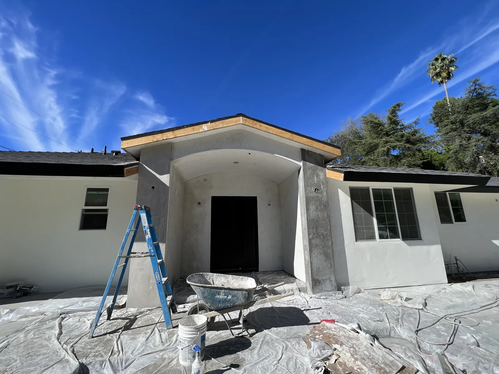 Front entry with fresh white stucco, a new arched portico, and a dark double door, with a ladder and wheelbarrow mid-work on the protected ground at PMK's addition-and-renovation project in La Cañada Flintridge.